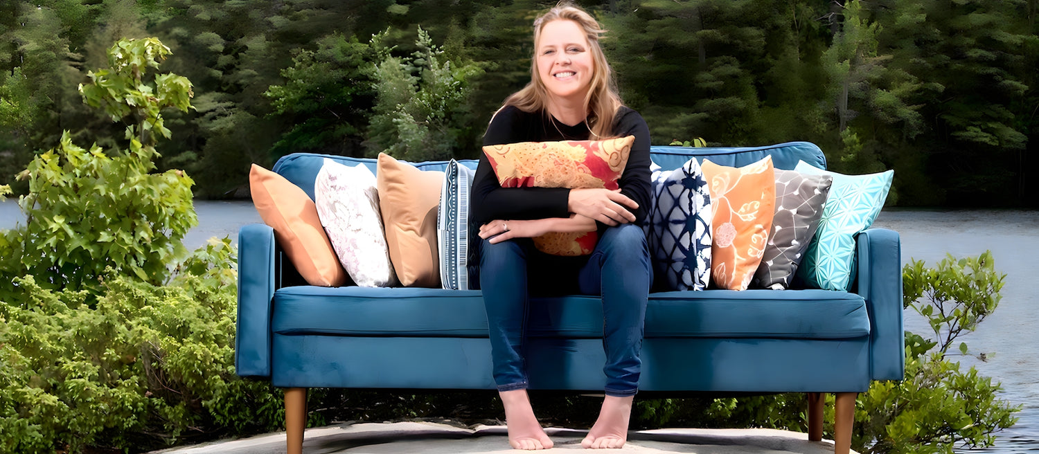 A person with long blonde hair is smiling while sitting barefoot on a blue outdoor sofa adorned with colorful patterned pillows. Lush green trees and a body of water are visible in the background.